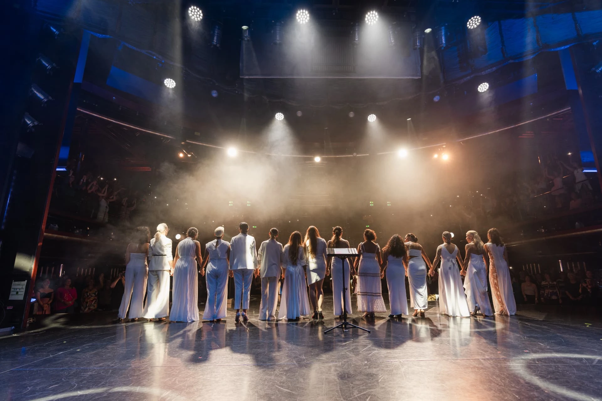 Le donne del movimento IAAWF, vestite di bianco, mano nella mano sul palco sotto le luci, davanti al pubblico.